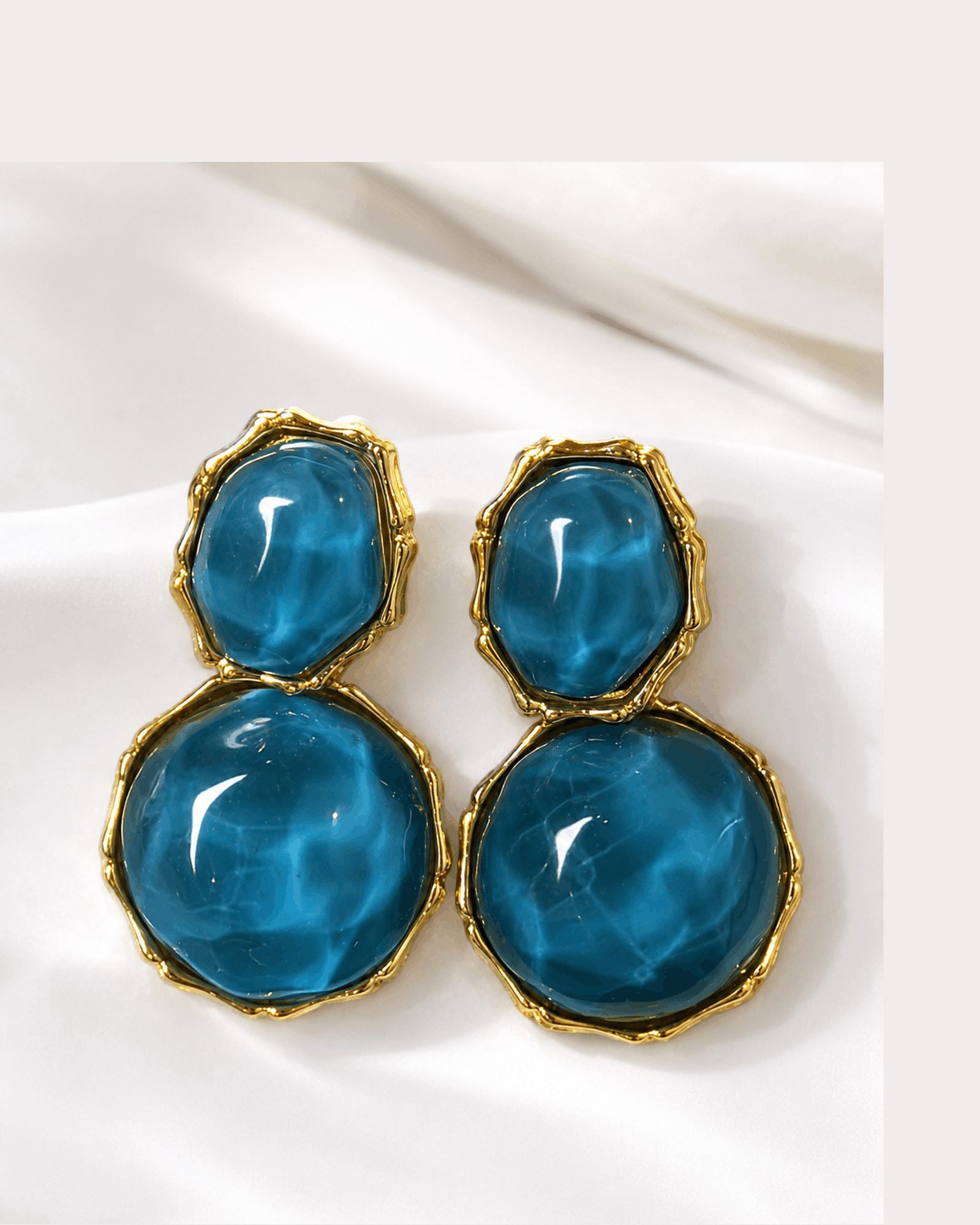 Azure Muse gold-tone earrings with blue resin stones photographed on ivory satin